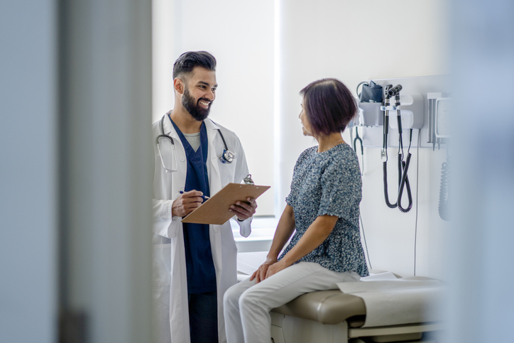 A young male doctor of Indian descent is at his office meeting with a senior female adult patient. His patient is seated on the examination table facing him. He is holding a clipboard and is smiling at his patient.